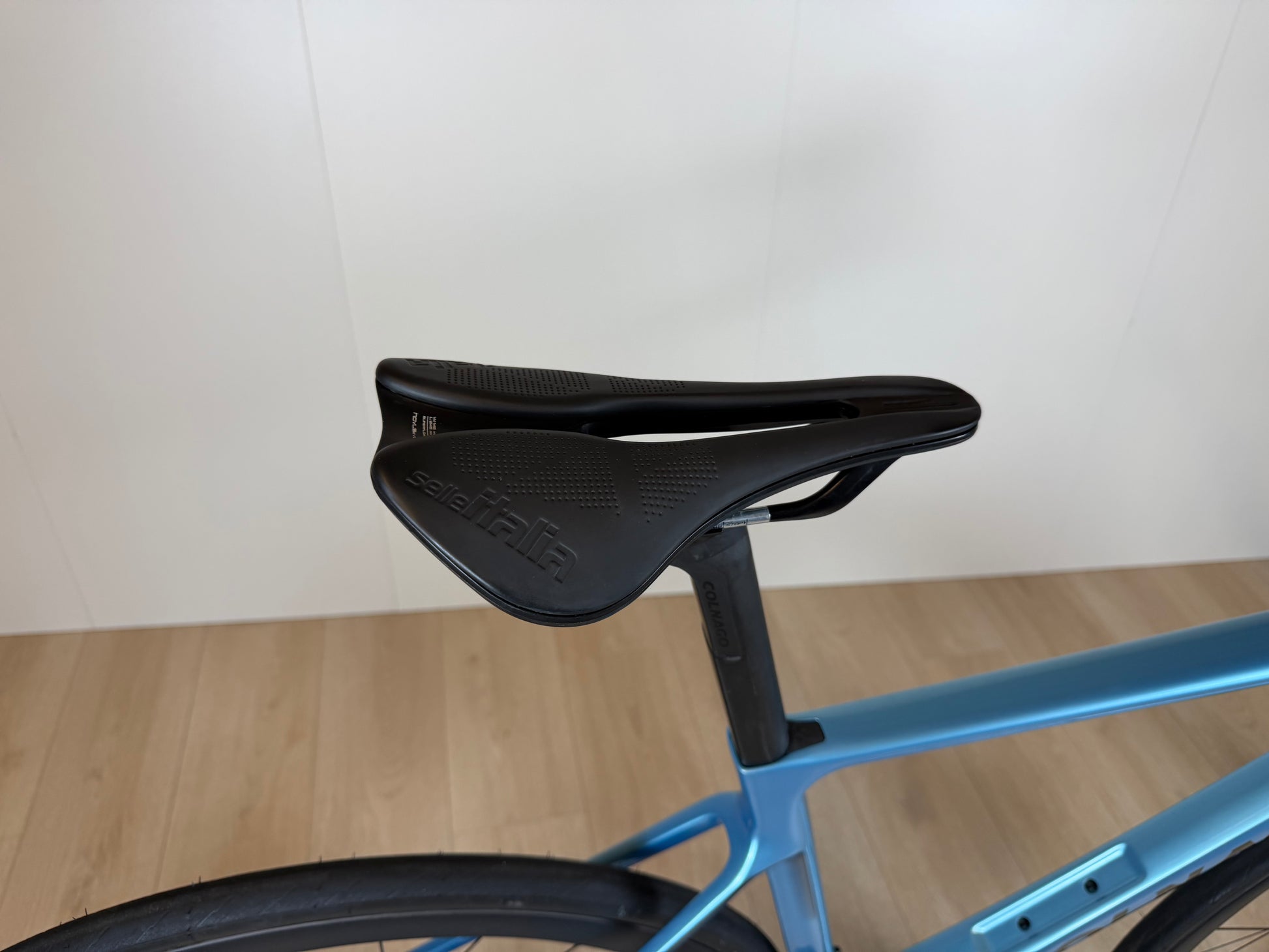 Zwart fietszadel op een blauwe Colnago V4 racefiets, zijaanzicht, lichte constructie, sportief ontwerp, binnenhuis op houten vloer.