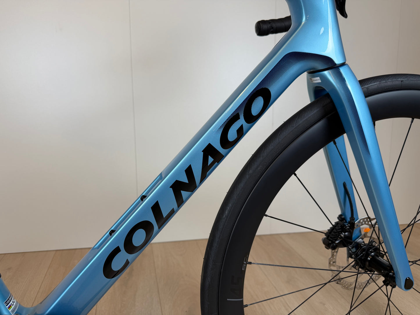 Zicht op een blauwe Colnago V4 racefiets met zwart logo, framemateriaal en voorvork, gefotografeerd van de zijkant op houten vloer.