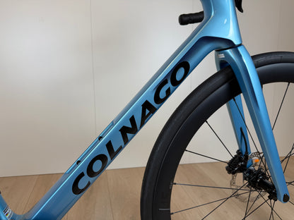 Zicht op een blauwe Colnago V4 racefiets met zwart logo, framemateriaal en voorvork, gefotografeerd van de zijkant op houten vloer.
