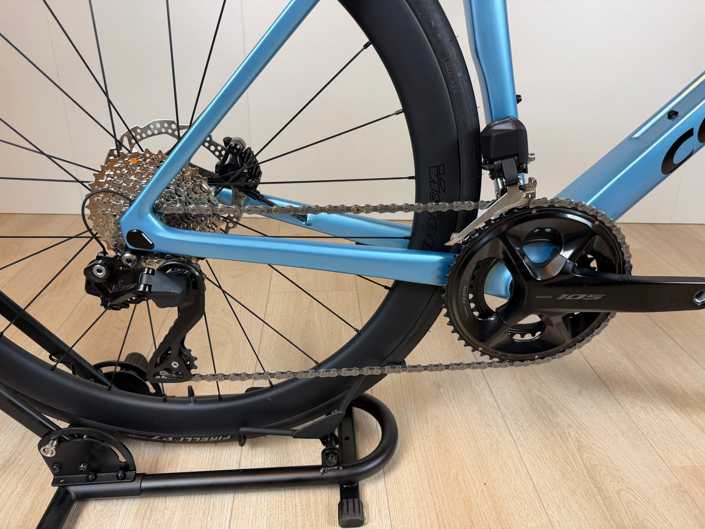 Zicht op de blauwe Colnago V4 racefiets met zwart crankstel en achterderailleur, gefotografeerd van de zijkant op een houten vloer.