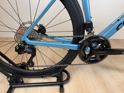 Zicht op de blauwe Colnago V4 racefiets met zwart crankstel en achterderailleur, gefotografeerd van de zijkant op een houten vloer.
